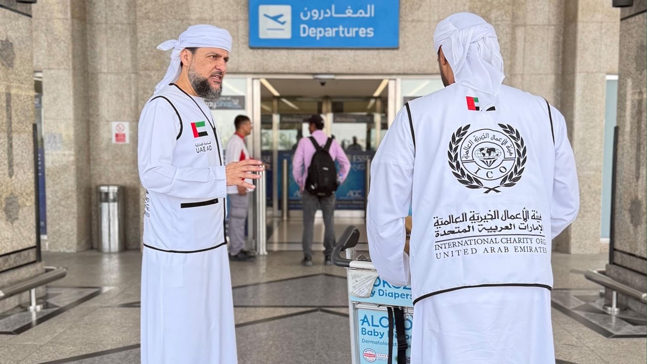 Two representatives of the International Charity Organization (ICO) from the United Arab Emirates stand at an airport departure terminal, preparing for a humanitarian mission. One team member is speaking while the other stands beside a luggage trolley, both wearing official charity vests displaying the UAE flag and organization logo.